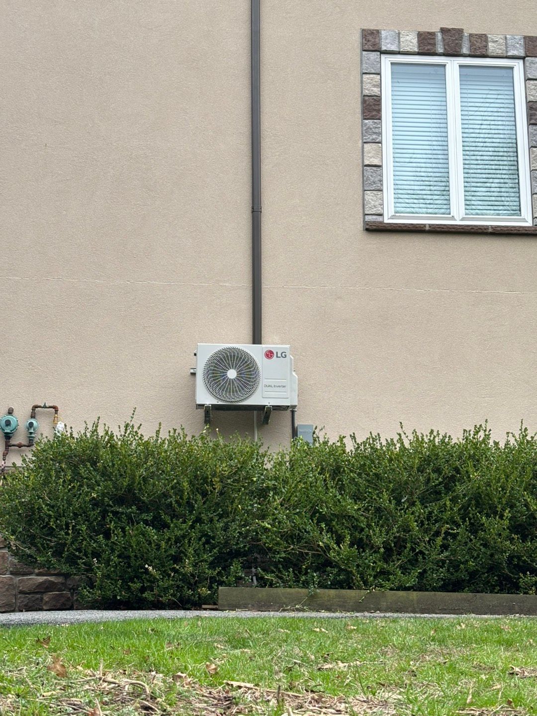 Outdoor wall with air-conditioning unit, window, and hedges in front of a beige building.