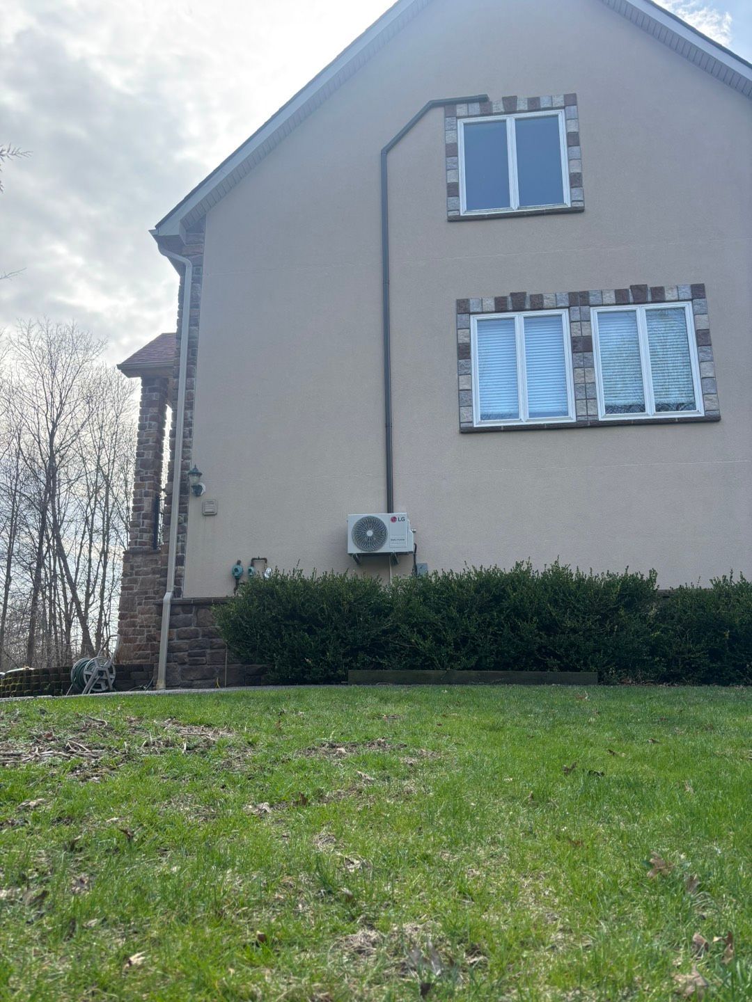 Beige house exterior with two windows, a wall-mounted air conditioner, and a grassy yard under a cloudy sky