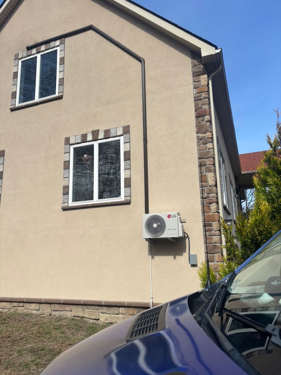 Exterior of a beige house with windows, stone trim, and a wall-mounted air conditioner under a blue sky