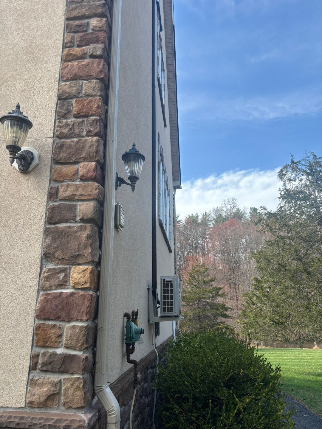 Side view of a house with stone corner trim, wall lanterns, and shrubs beside a grassy yard.