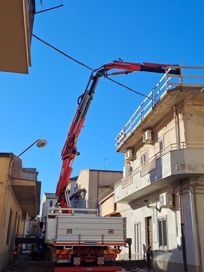 Un camion con gru rosso solleva materiali sul tetto di una stretta via cittadina sotto un cielo azzurro e limpido.