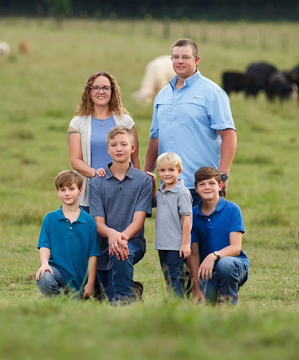 Family of six posing in a grassy field with cattle. Smiling faces, blue and gray clothing.