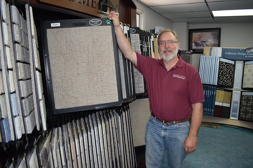 A man in a maroon shirt is standing in front of a display of carpets