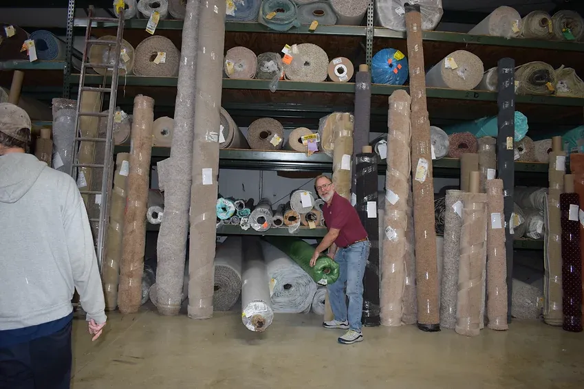 A man is standing in a warehouse full of rolls of carpet