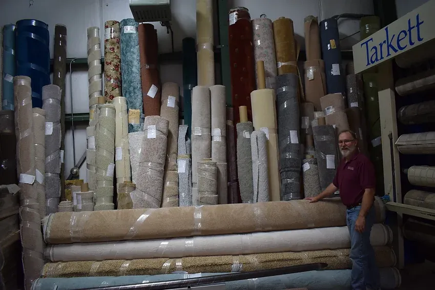 A man is standing in front of a large stack of carpets in a store called tarkett