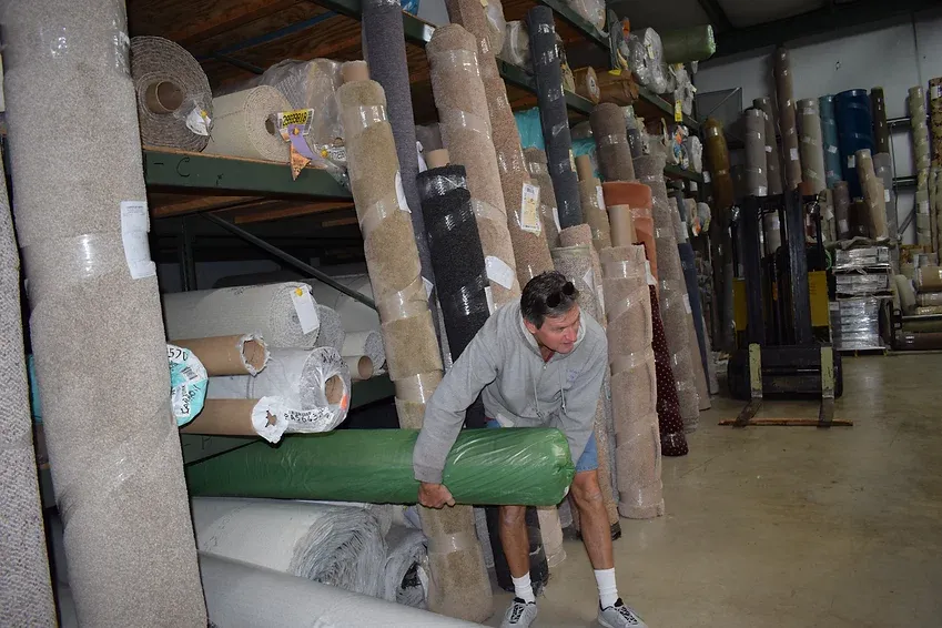A man is carrying a green roll of carpet in a warehouse.