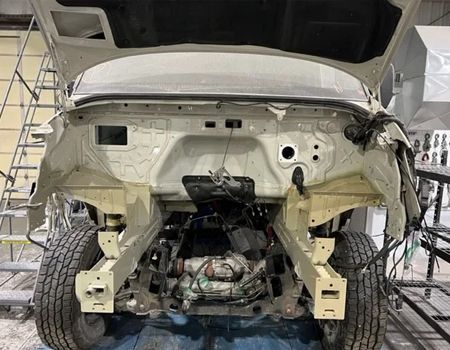 A white vehicle frame stripped of its engine and hood, resting on tires in an indoor workshop setting.
