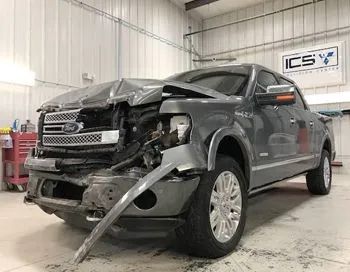 Damaged gray Ford F-150 pickup truck inside a repair shop. Front end heavily damaged from a collision.