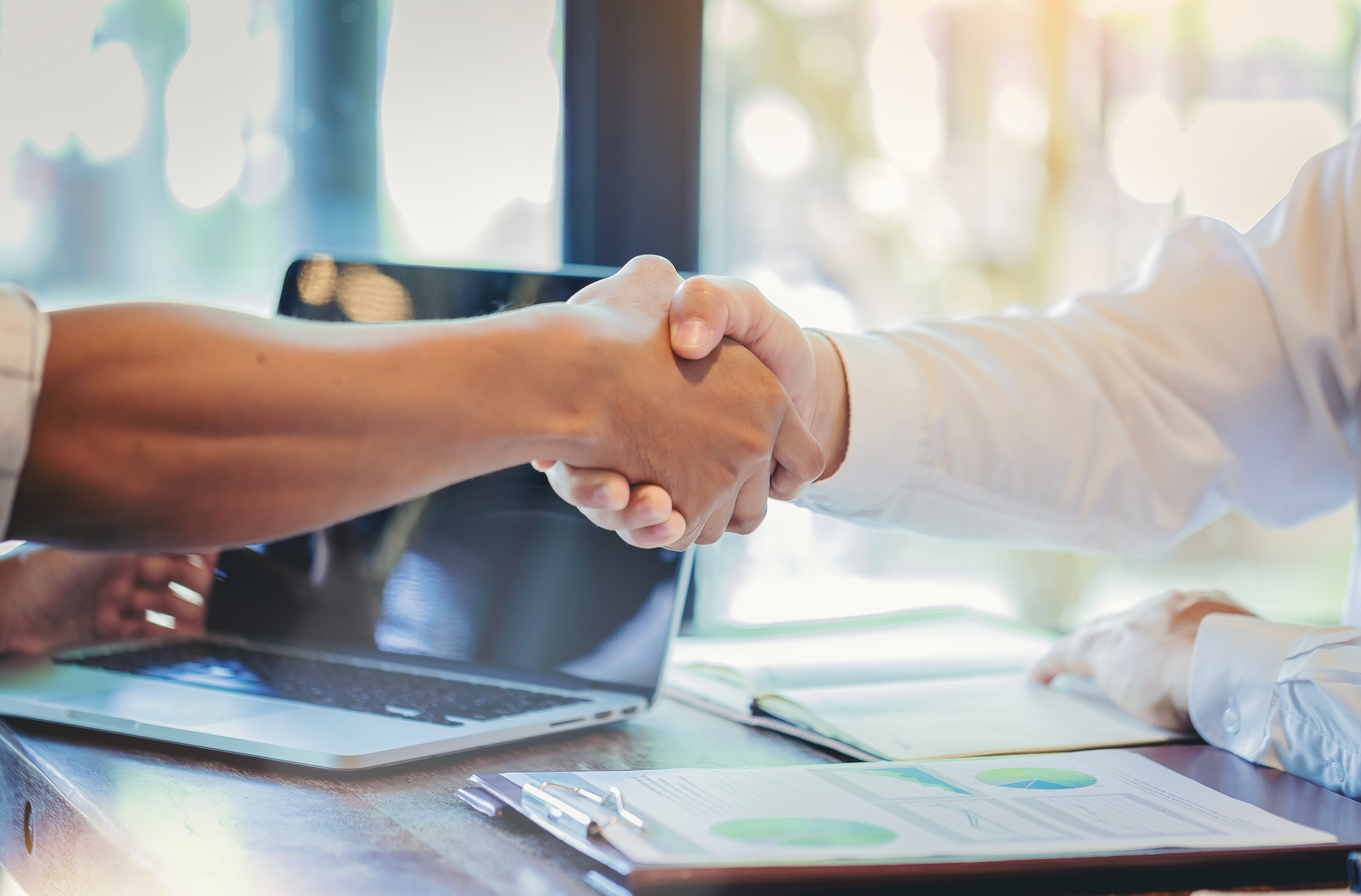 Two people are shaking hands over a table with a laptop.