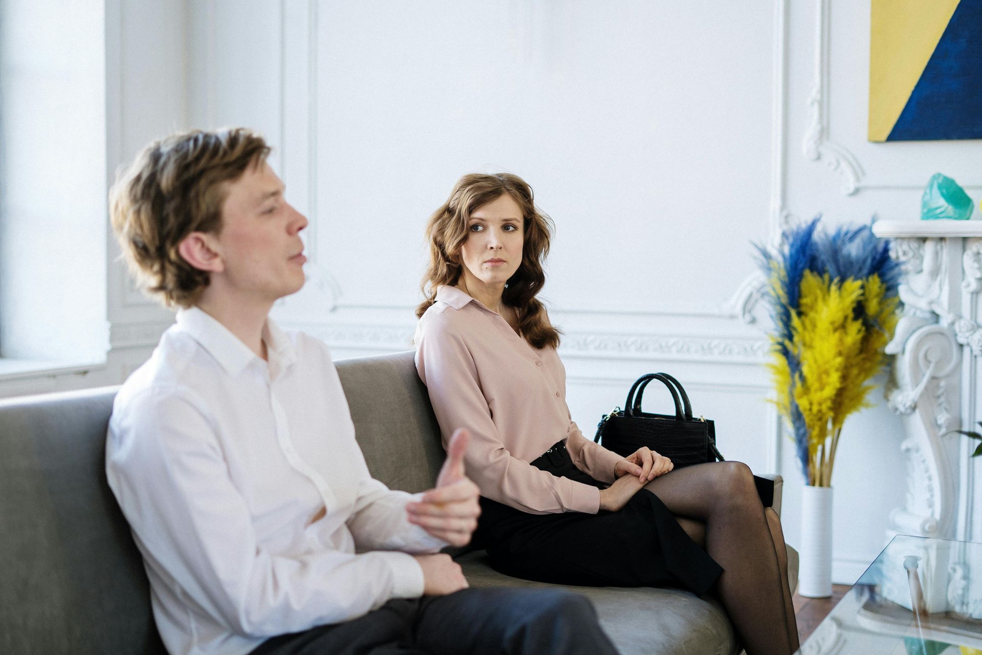 Two people seated on a gray couch in a bright room, one holding a black handbag, with blue flowers nearby