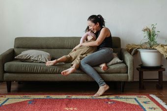 Person hugging a child on a green sofa in a bright living room, with a rug and potted plant nearby.