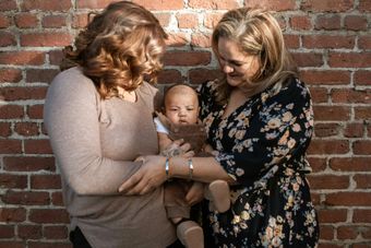 Two women holding a baby in front of a brick wall, smiling and looking at the child.