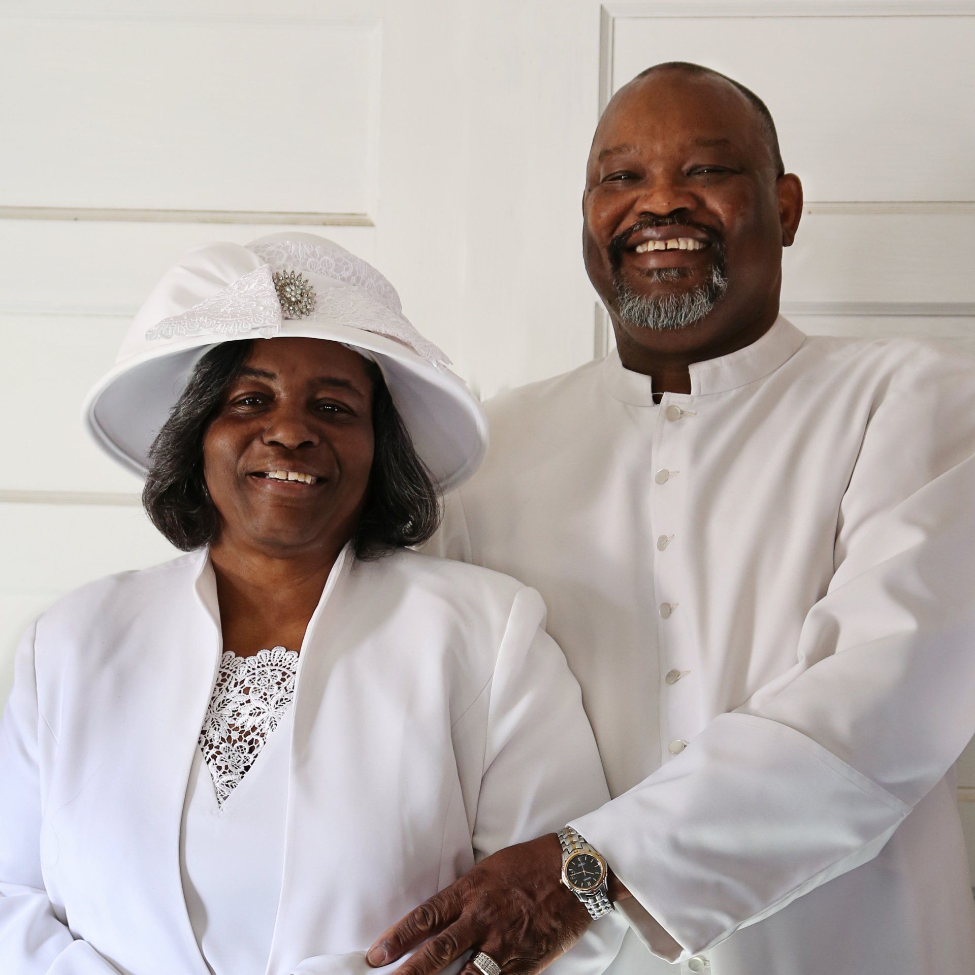 Couple in white outfits smiling, posed indoors. Woman wears a hat, man has a watch.