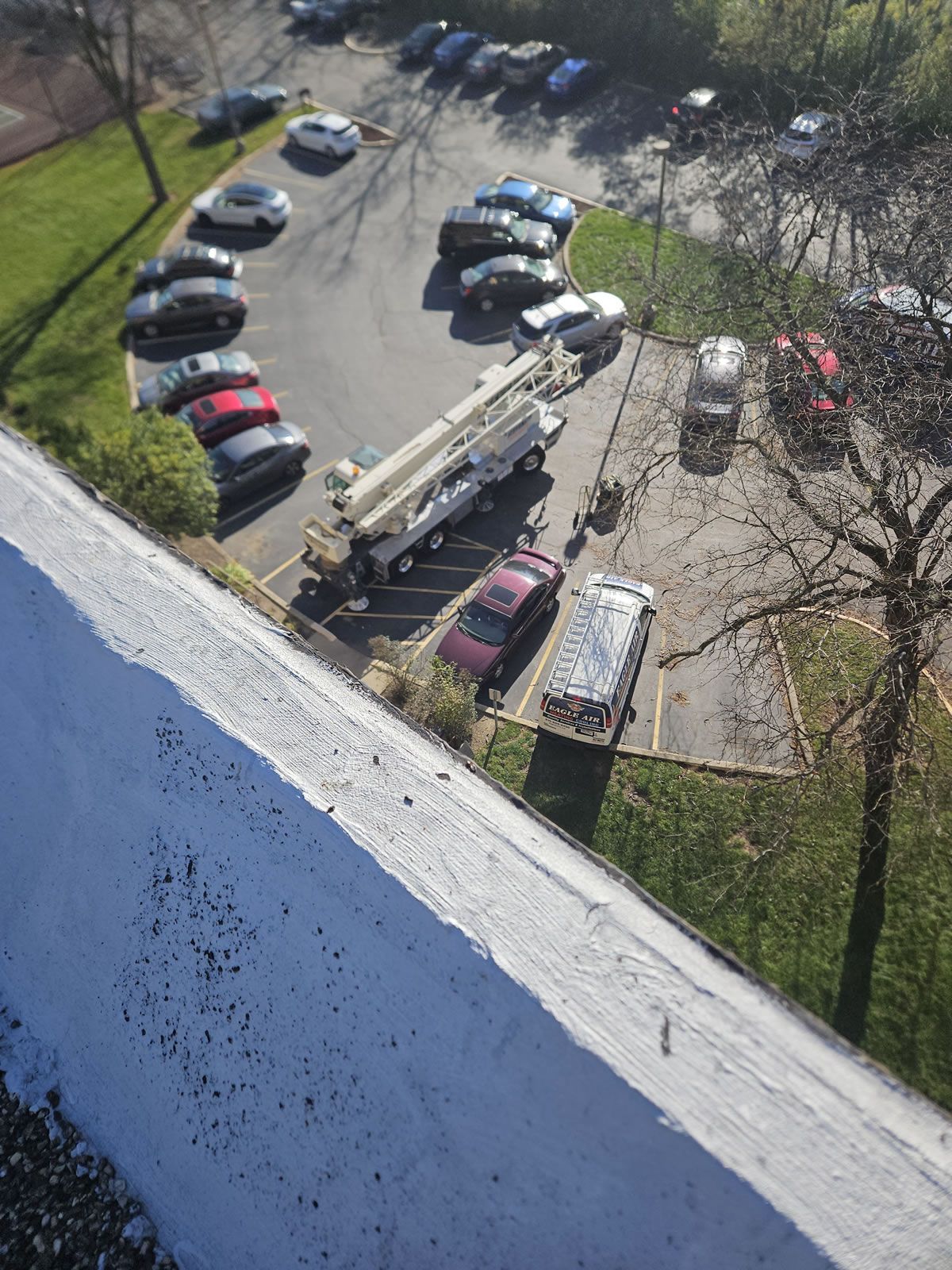 A parking lot with cars and a large crane. Bright sunlight, some green grass, and a rooftop in the foreground.