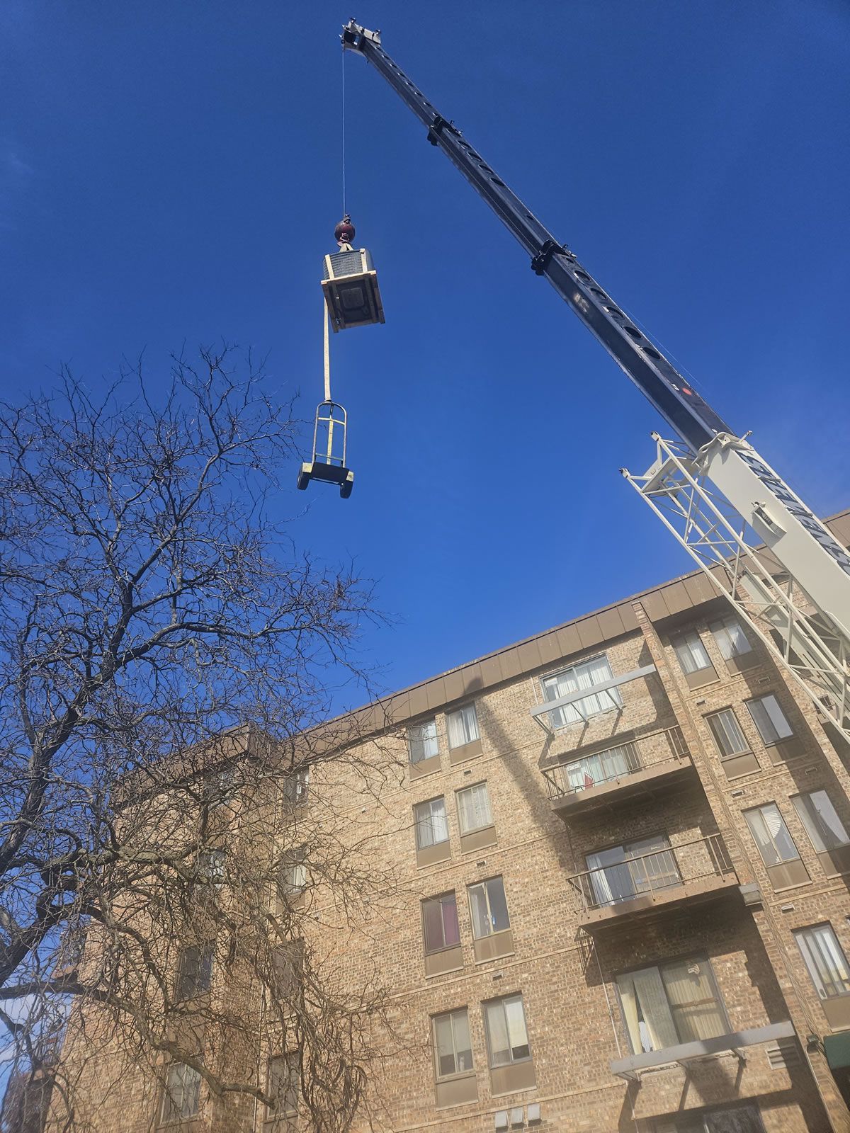Crane lifting equipment to the rooftop of a brick apartment building on a sunny day.