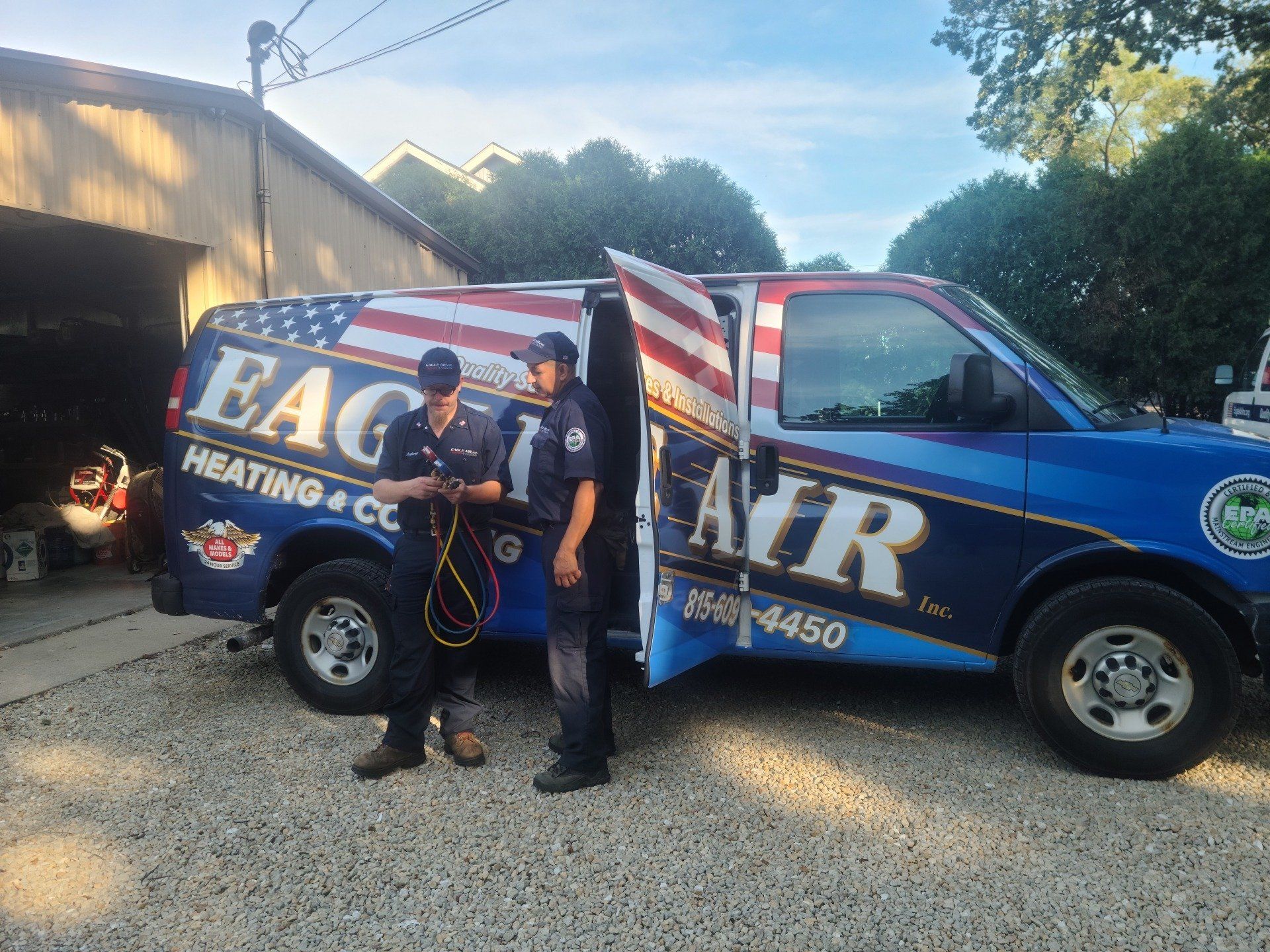 Two men in uniform beside a blue HVAC van with American flag design; in a driveway.