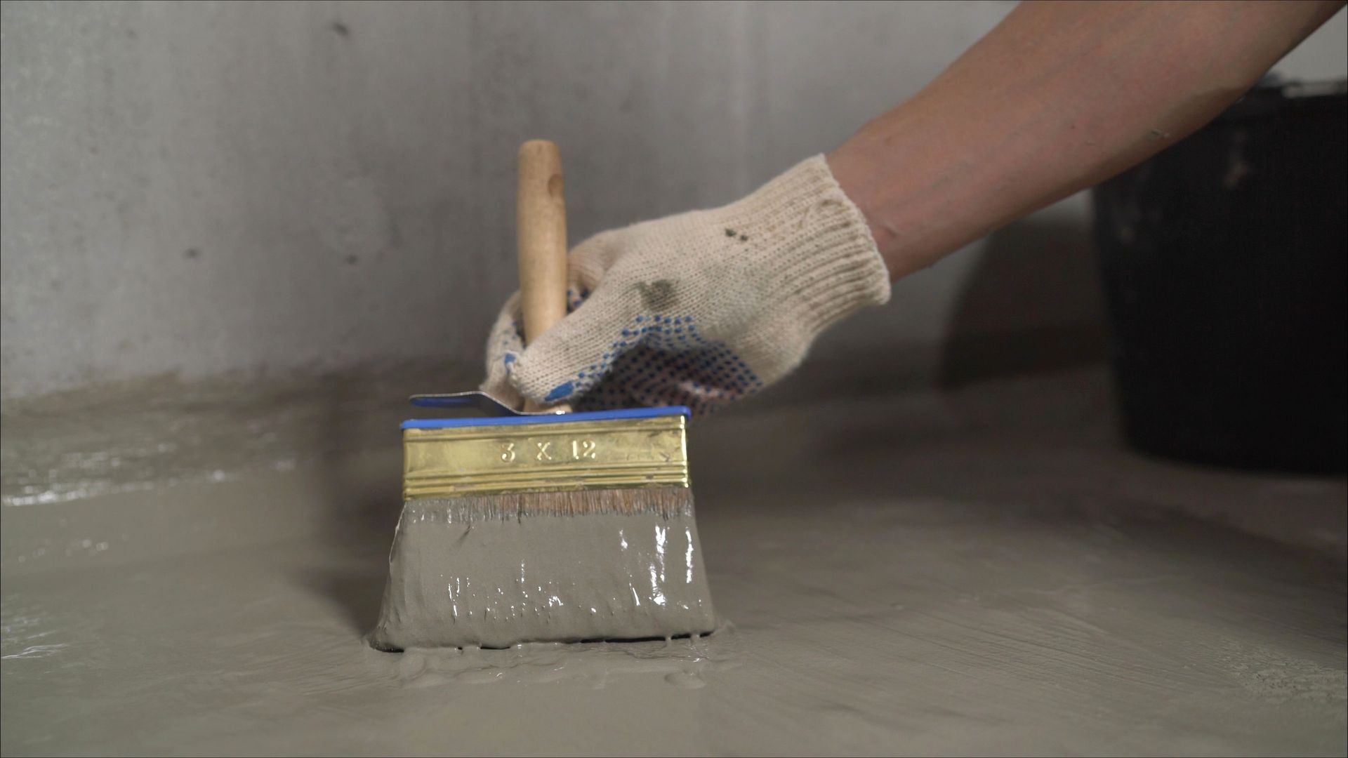 Gloved hand brushes gray waterproof coating onto a concrete basement floor.