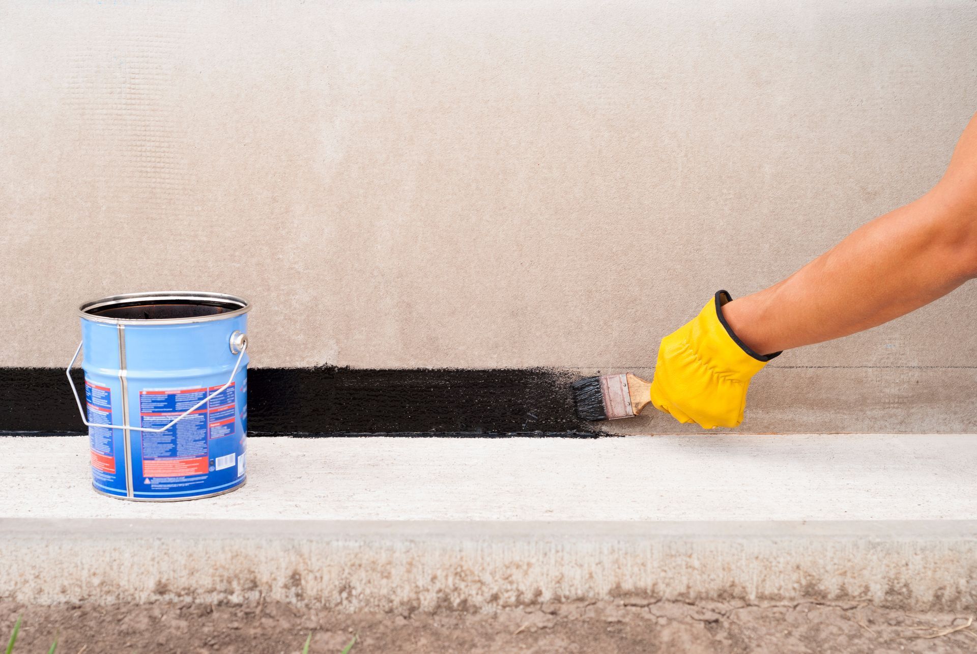 Hand in a yellow glove brushing waterproof coating along a concrete wall base. Hand in a yellow glove brushing waterproof coating along a concrete wall base.