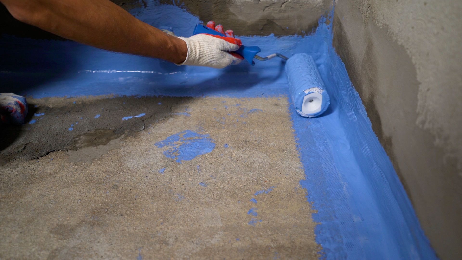 Worker applies blue waterproof coating with a paint roller along a concrete floor edge.