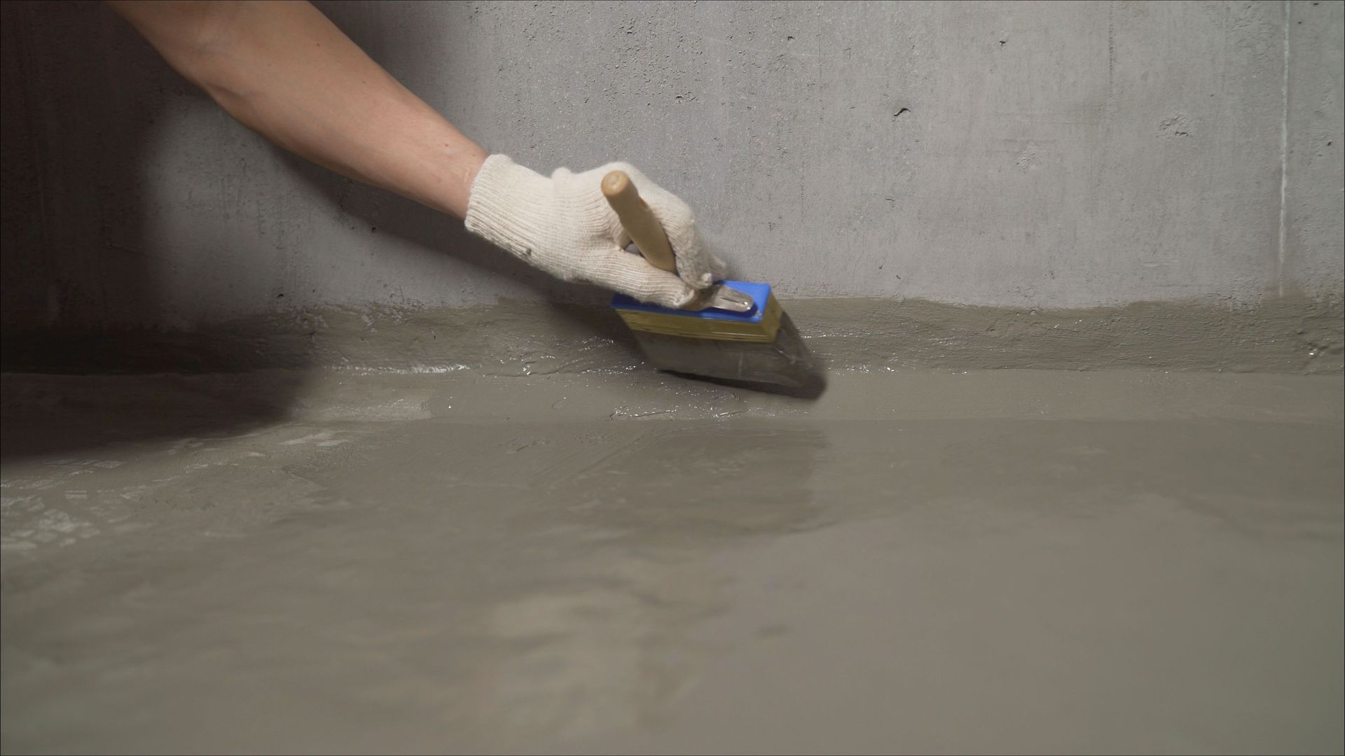 Gloved hand applies gray waterproof sealant along a concrete wall and floor joint.
