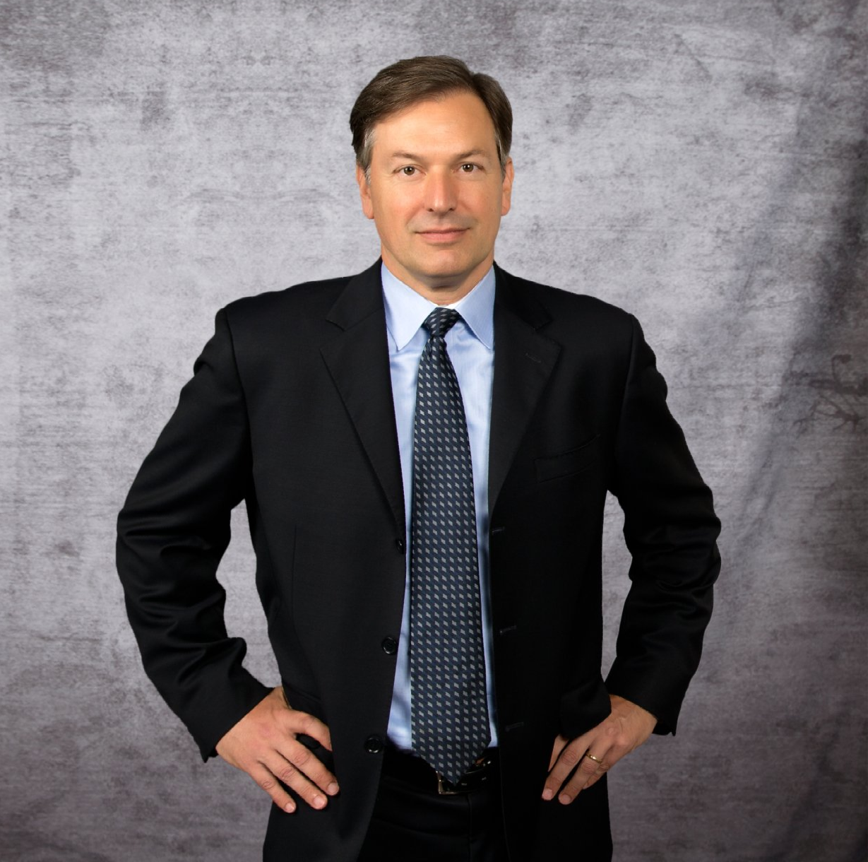 Man in a dark suit and tie standing against a gray studio backdrop, hands on hips.