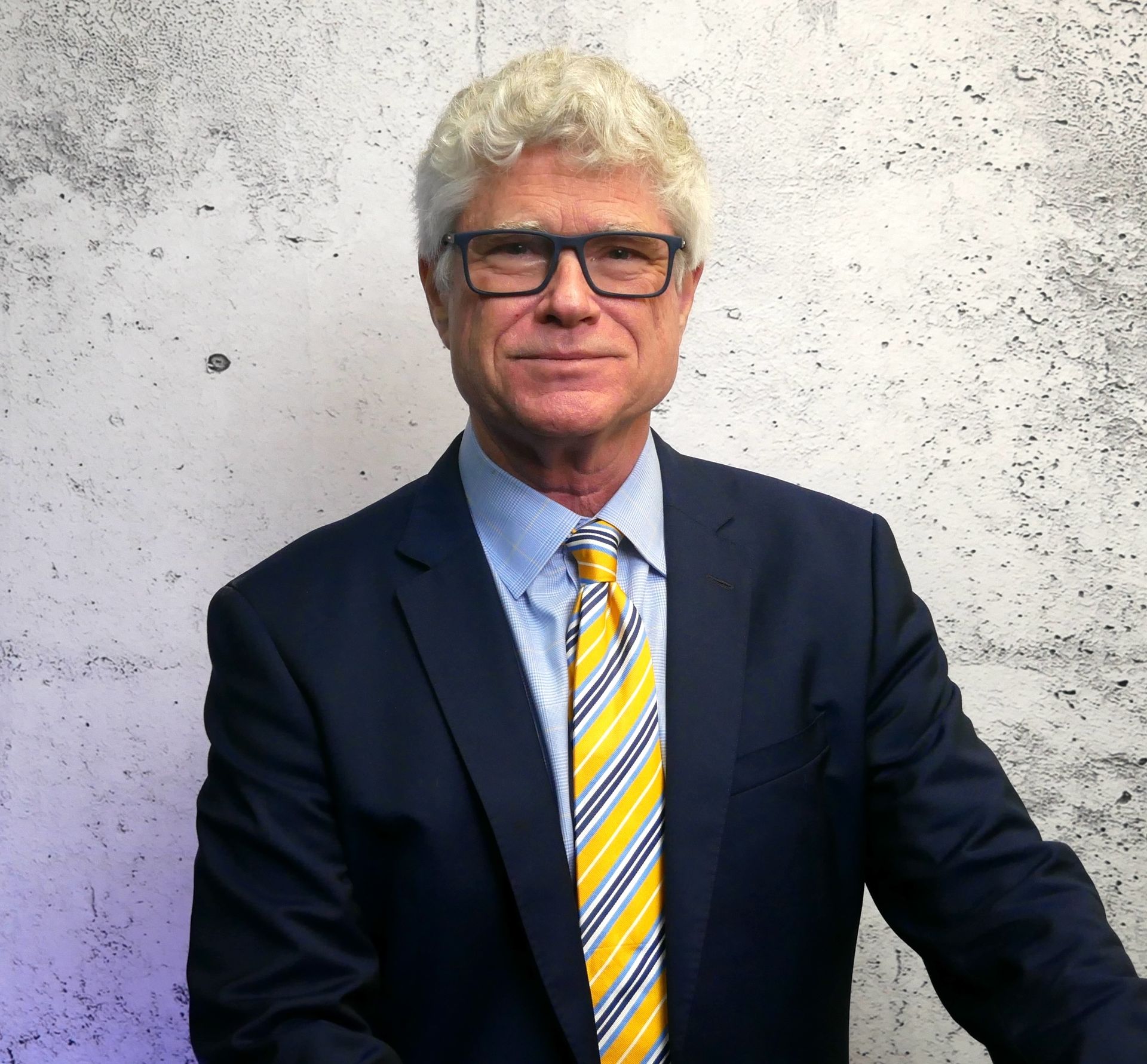 Man in a navy suit and striped tie standing against a speckled gray wall.