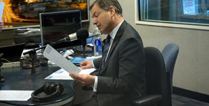 Man in a suit reading papers at a desk with a microphone in a radio studio