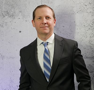 Man in a black suit and blue striped tie standing against a gray textured wall