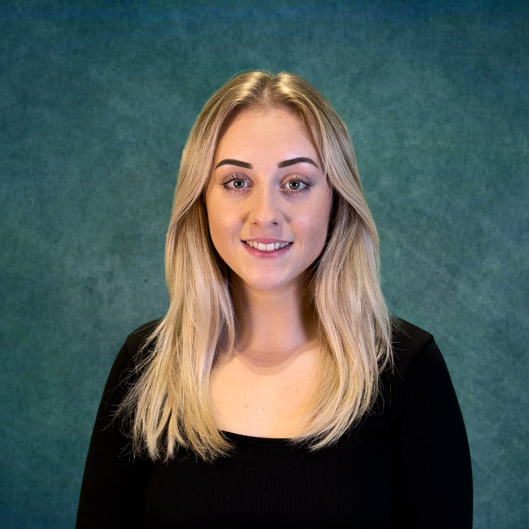 Blonde woman with long hair smiles, wearing a black shirt, against a teal background.