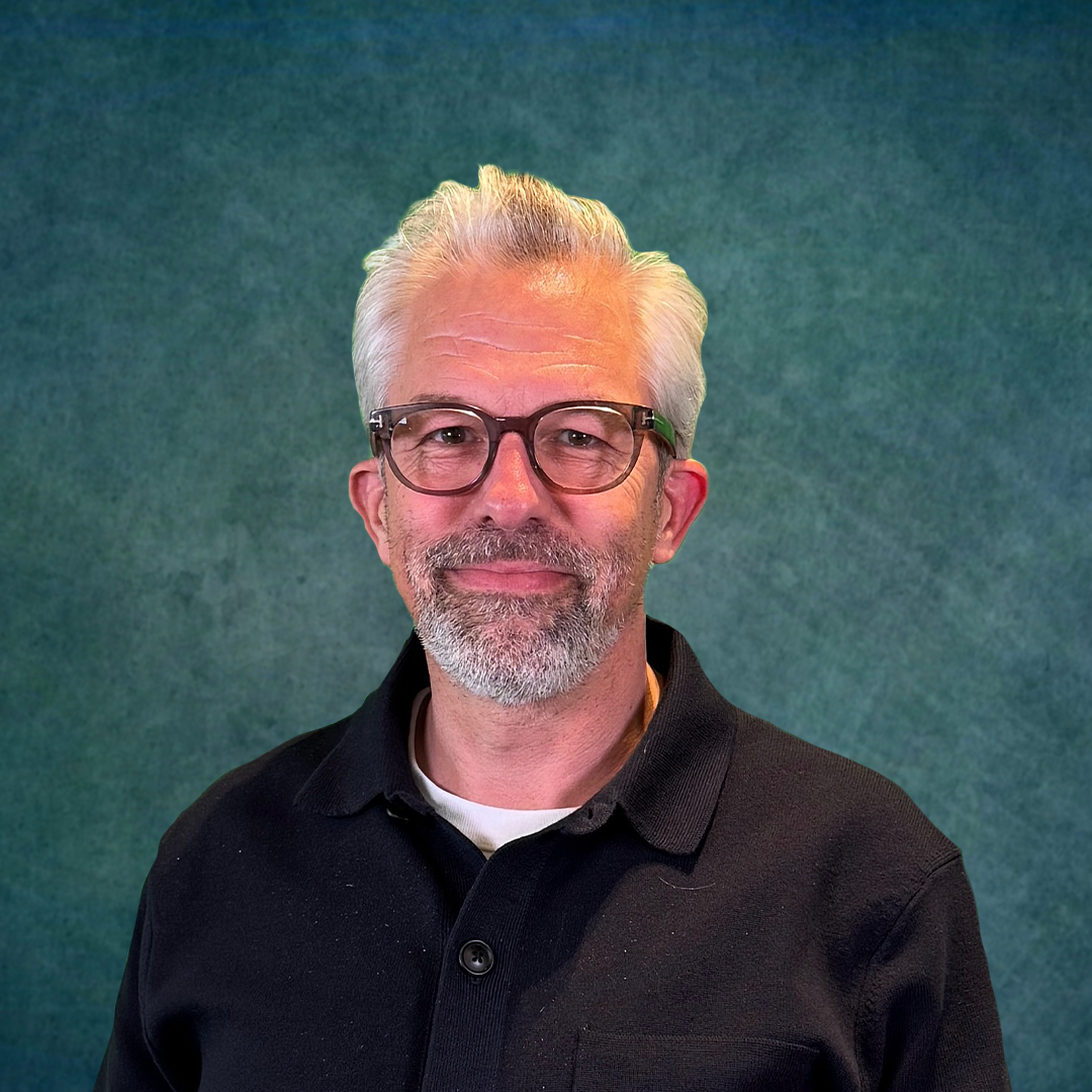 A man with glasses and a beard is standing in front of a colorful brick wall.