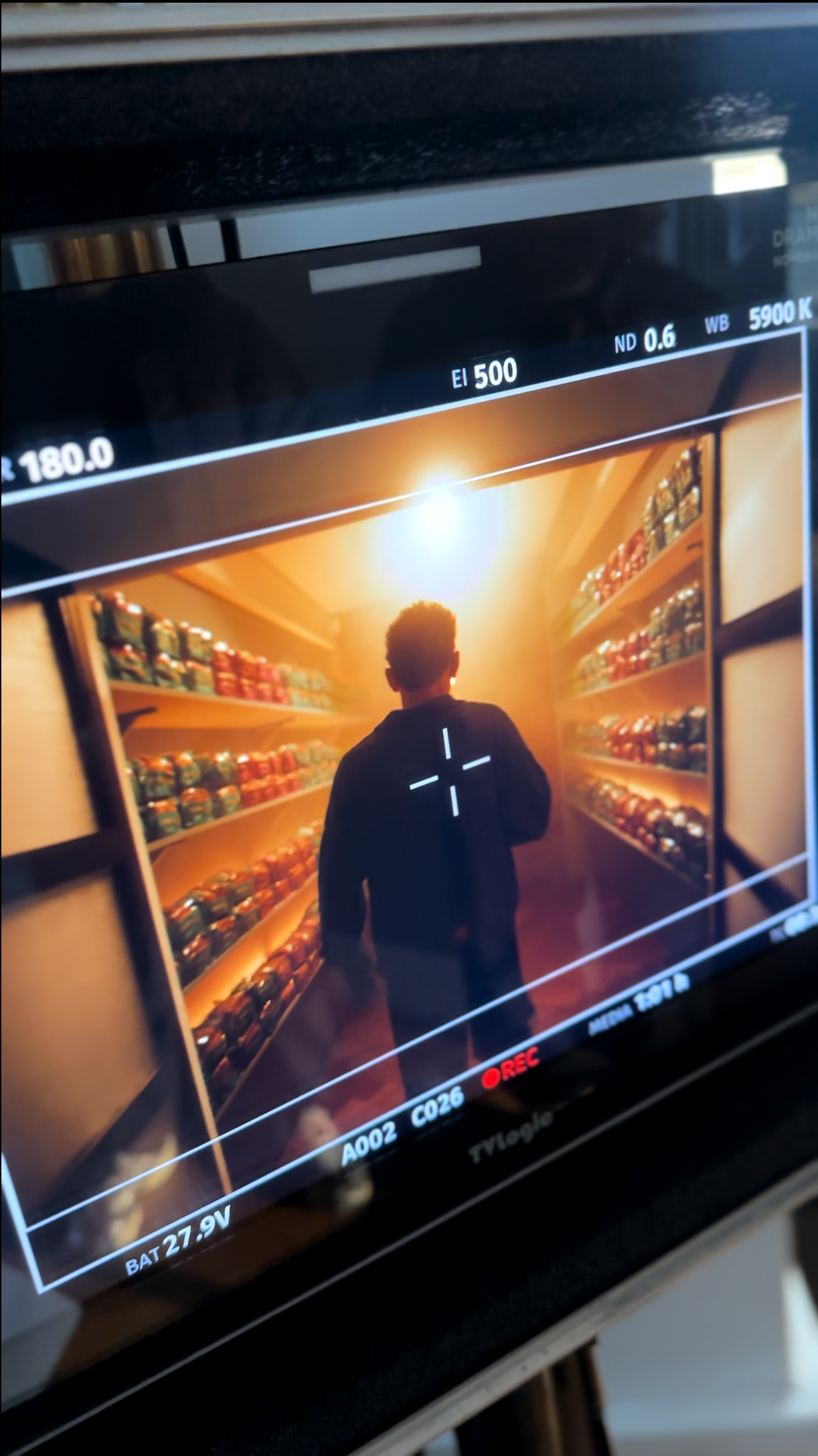 On-set monitor view: person walking toward bright light at the end of a shelf-lined corridor, possibly a wine cellar.