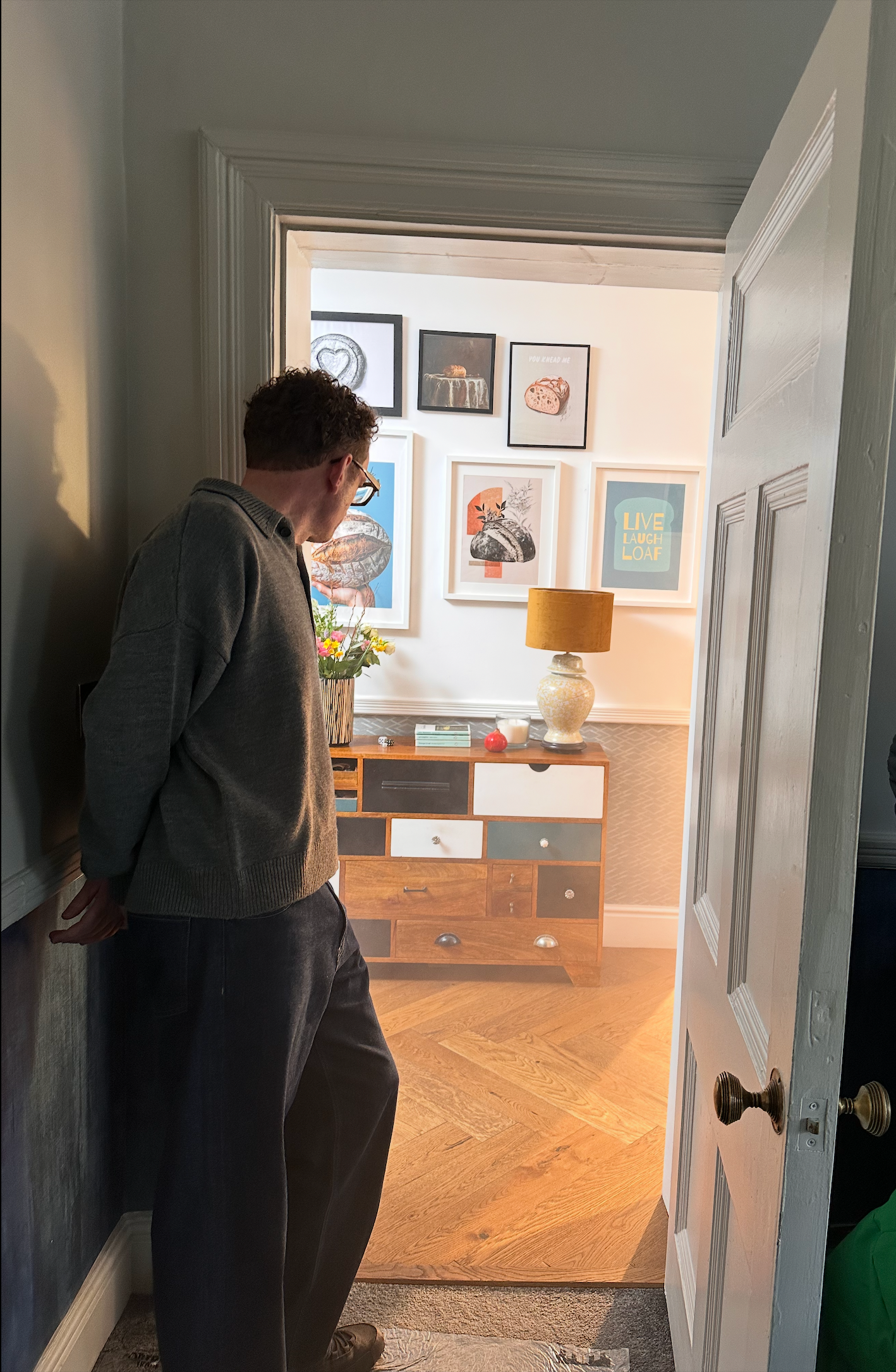 Man peers into a room with framed artwork, a colorful cabinet, and a glowing lamp.
