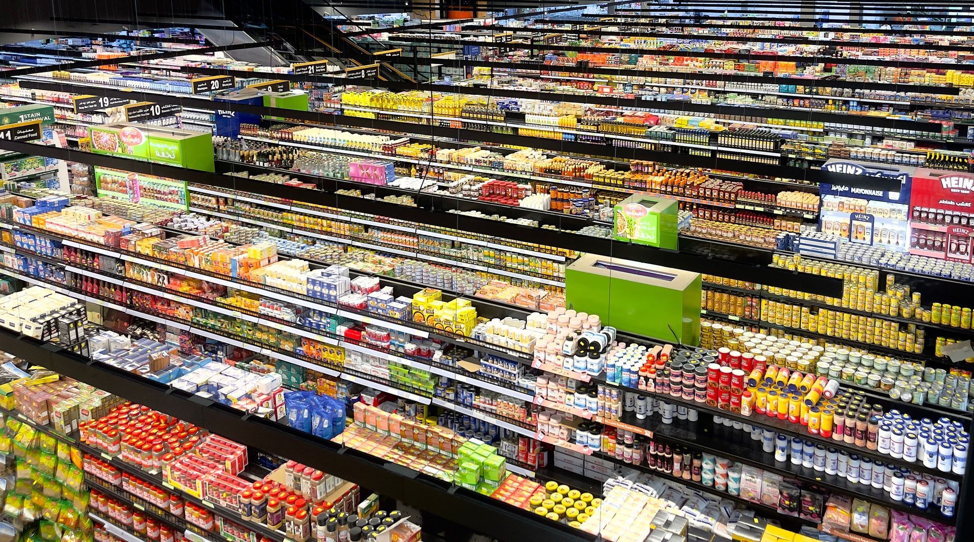 Aisle of a brightly lit grocery store with shelves of colorful packaged goods and a checkout sign ahead