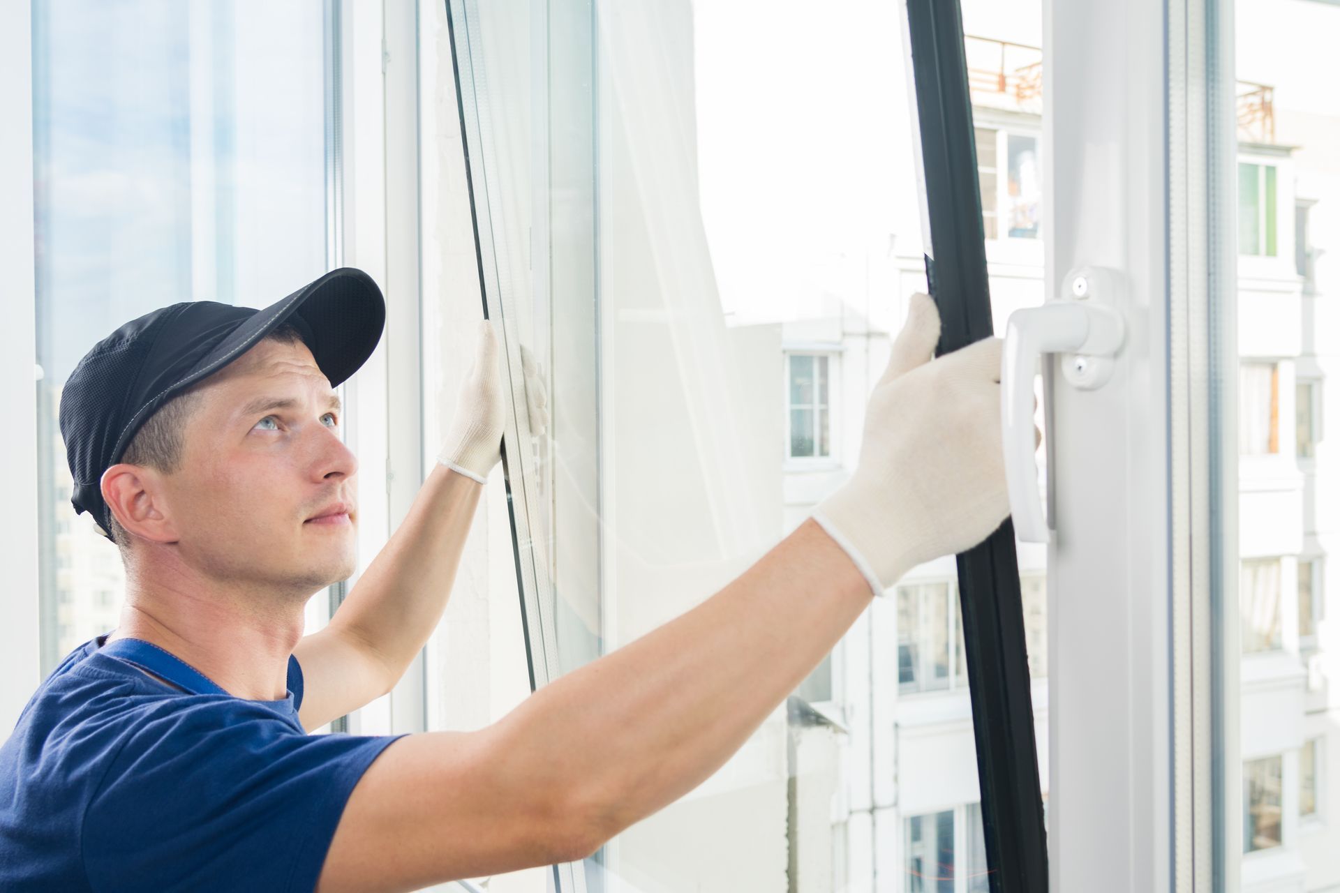 A person wearing a blue shirt, black cap, and gloves repairs the rubber seal on a white window frame.