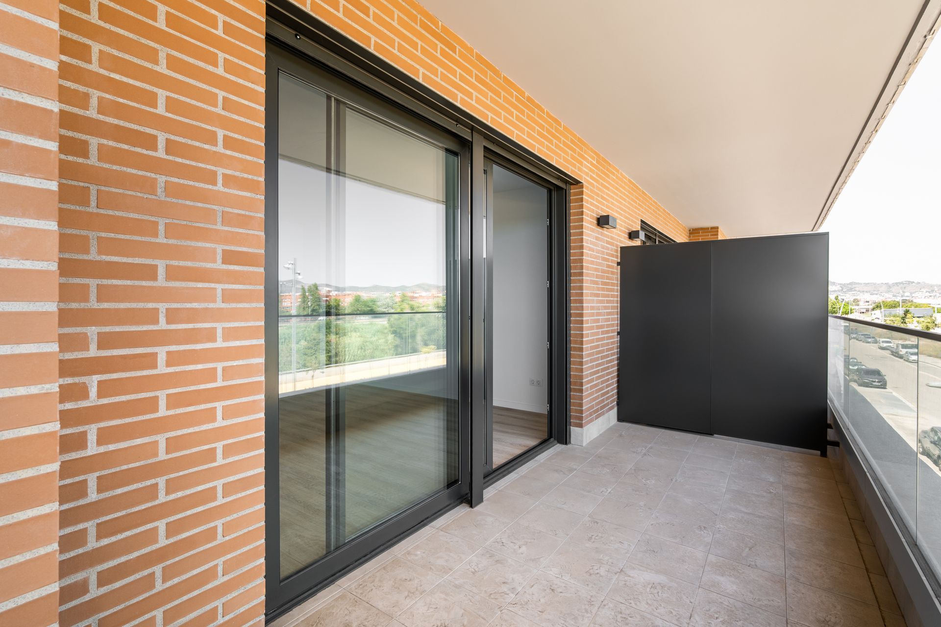 A sunny balcony with light-colored stone flooring, brick walls, a sliding glass door, and a glass railing.