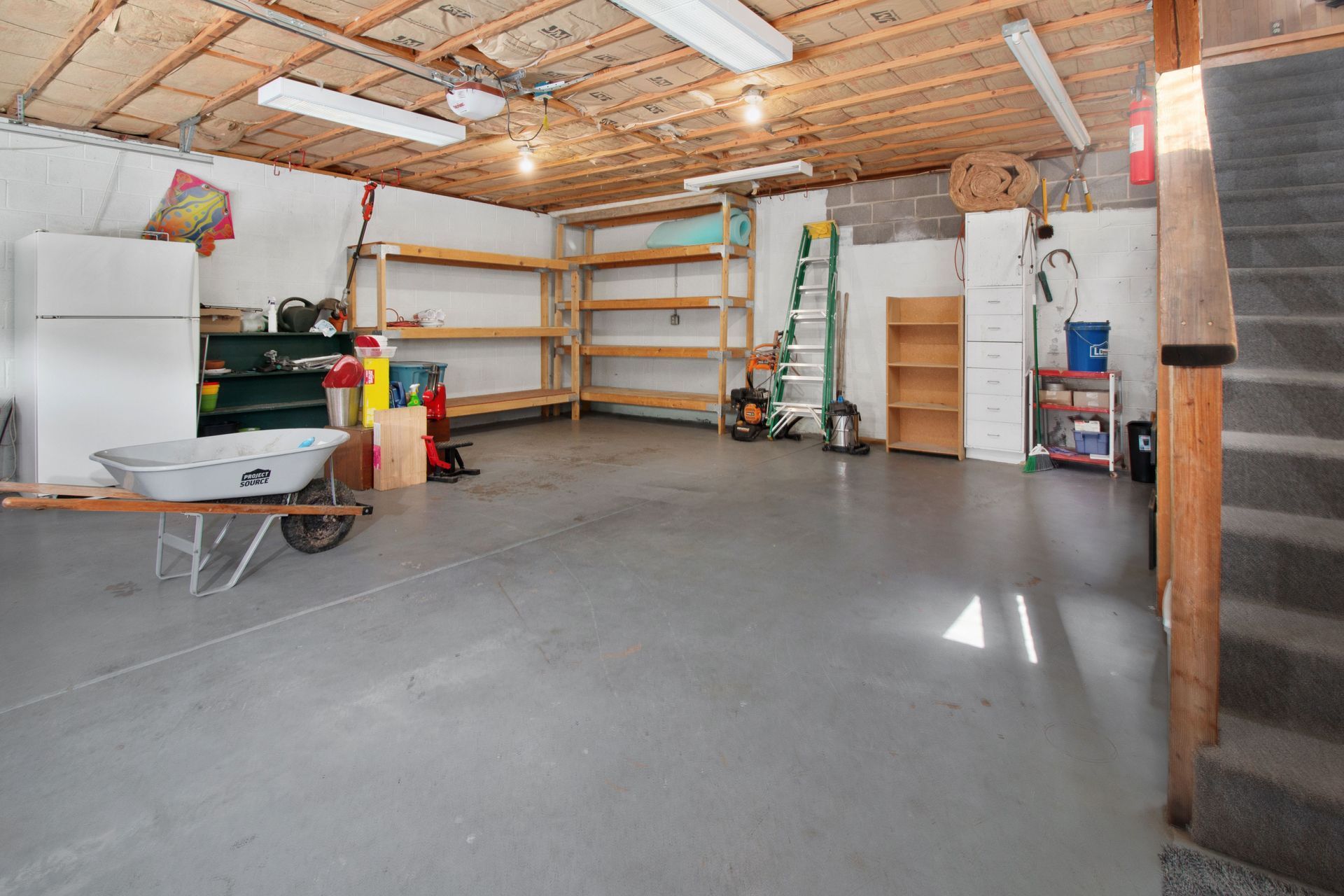 Empty garage with concrete floor, shelving, ladder, and stored household items under exposed wooden beams.