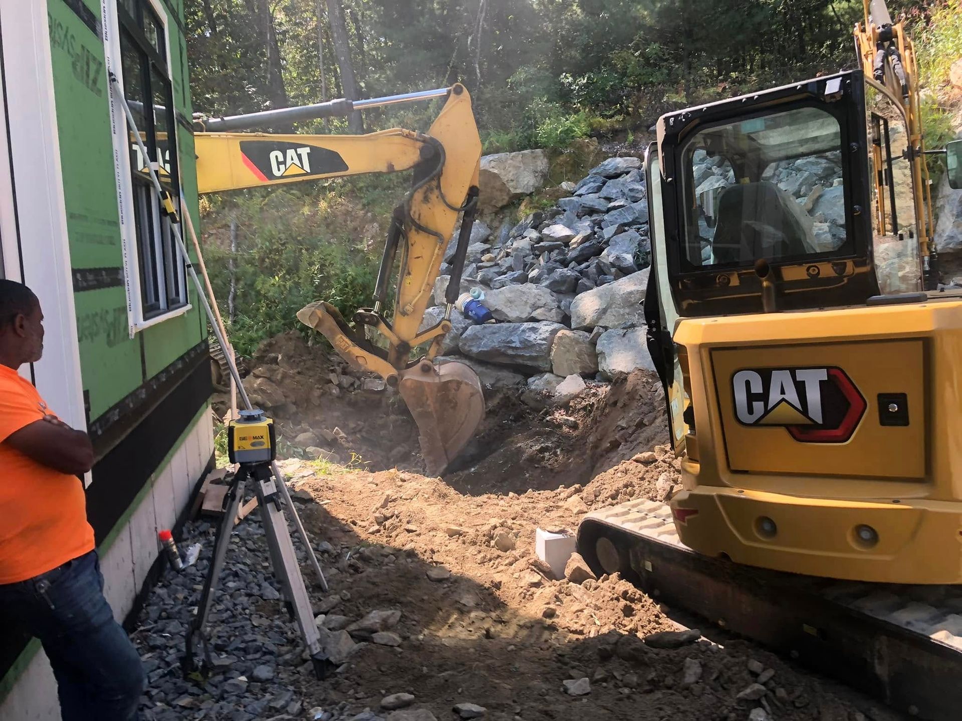 Construction site: excavator digging near a building, with a worker observing. Yellow equipment, sunny day.