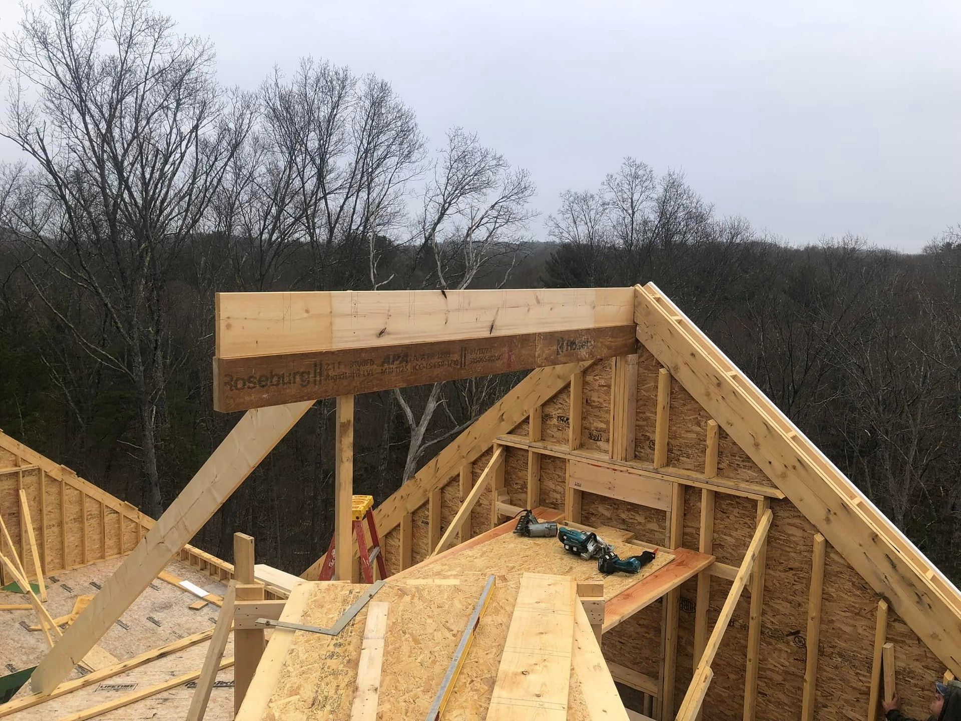 Framing of a roof in progress, with wooden trusses and supporting beams, outdoors against a forest backdrop.