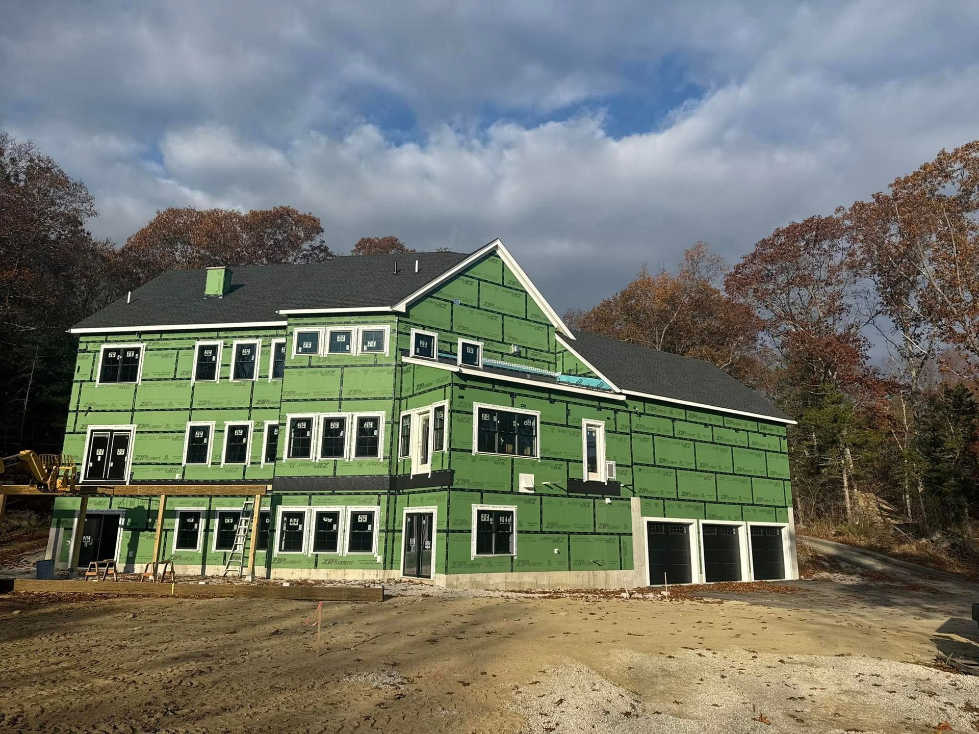 New house under construction, green sheathing, windows, black roof, dirt ground, trees, and cloudy sky.