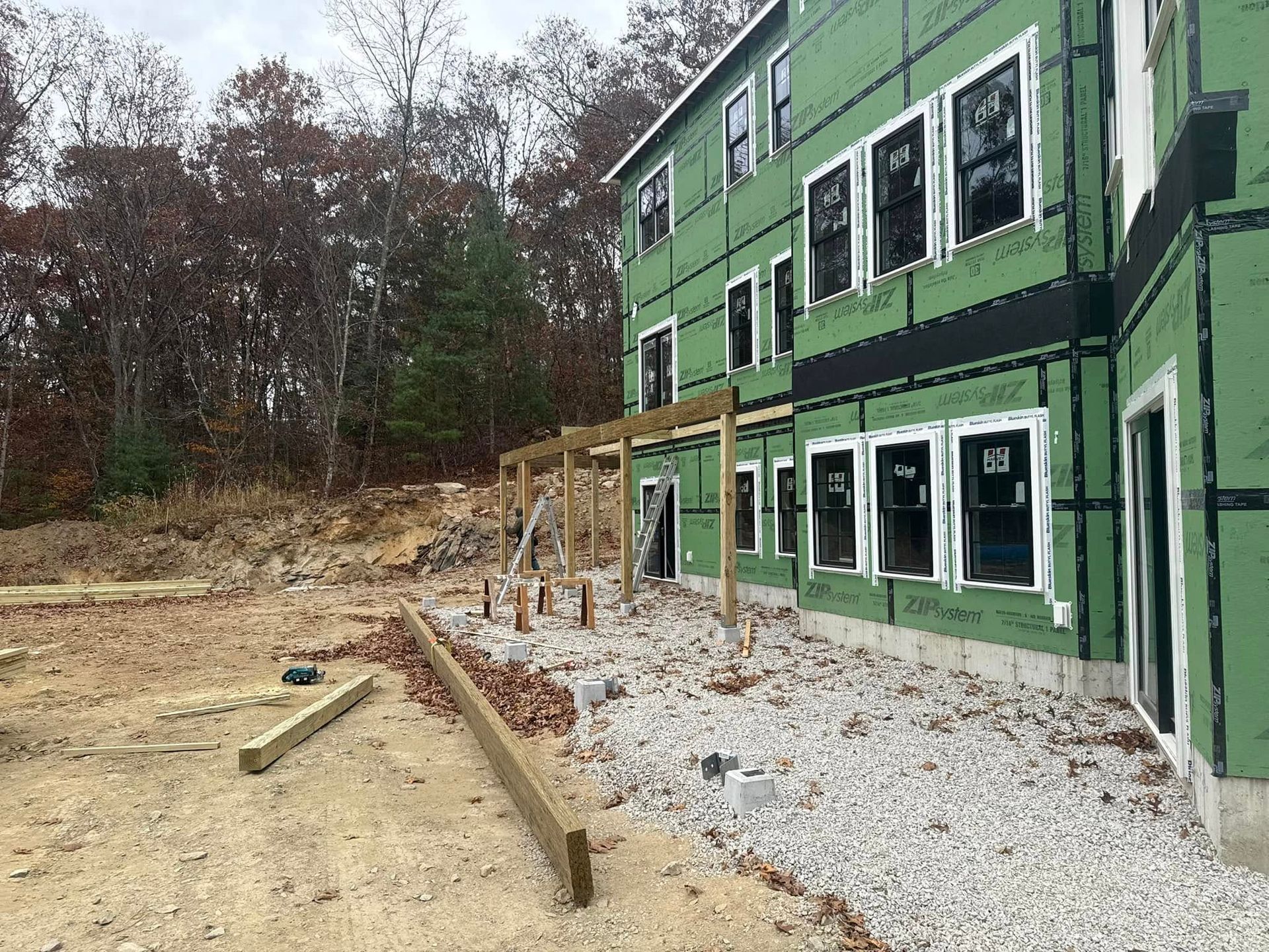 Construction site: green-wrapped building with wooden supports for a porch, gravel ground.
