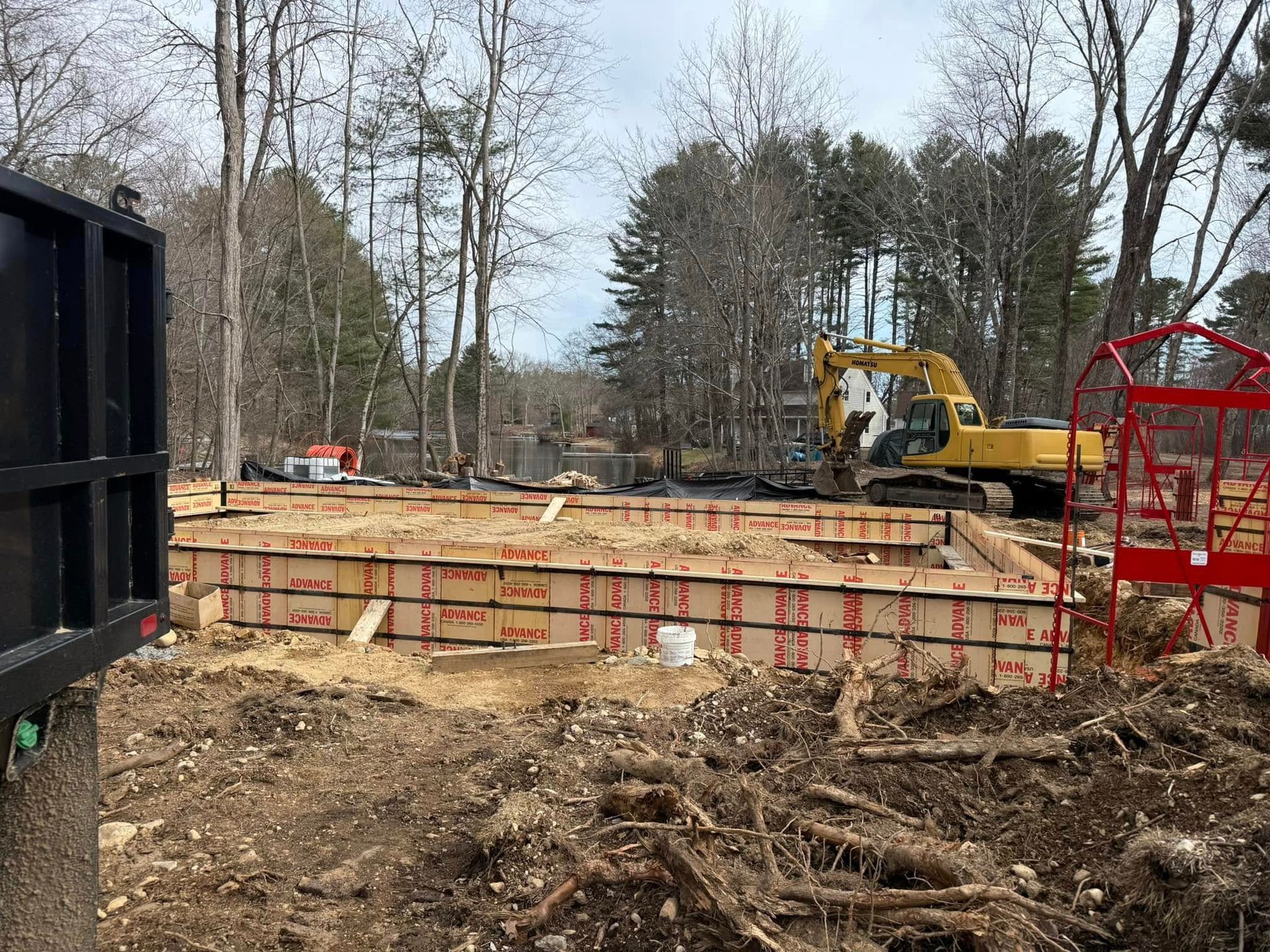 Construction site with foundation forms, excavator, and surrounding trees.