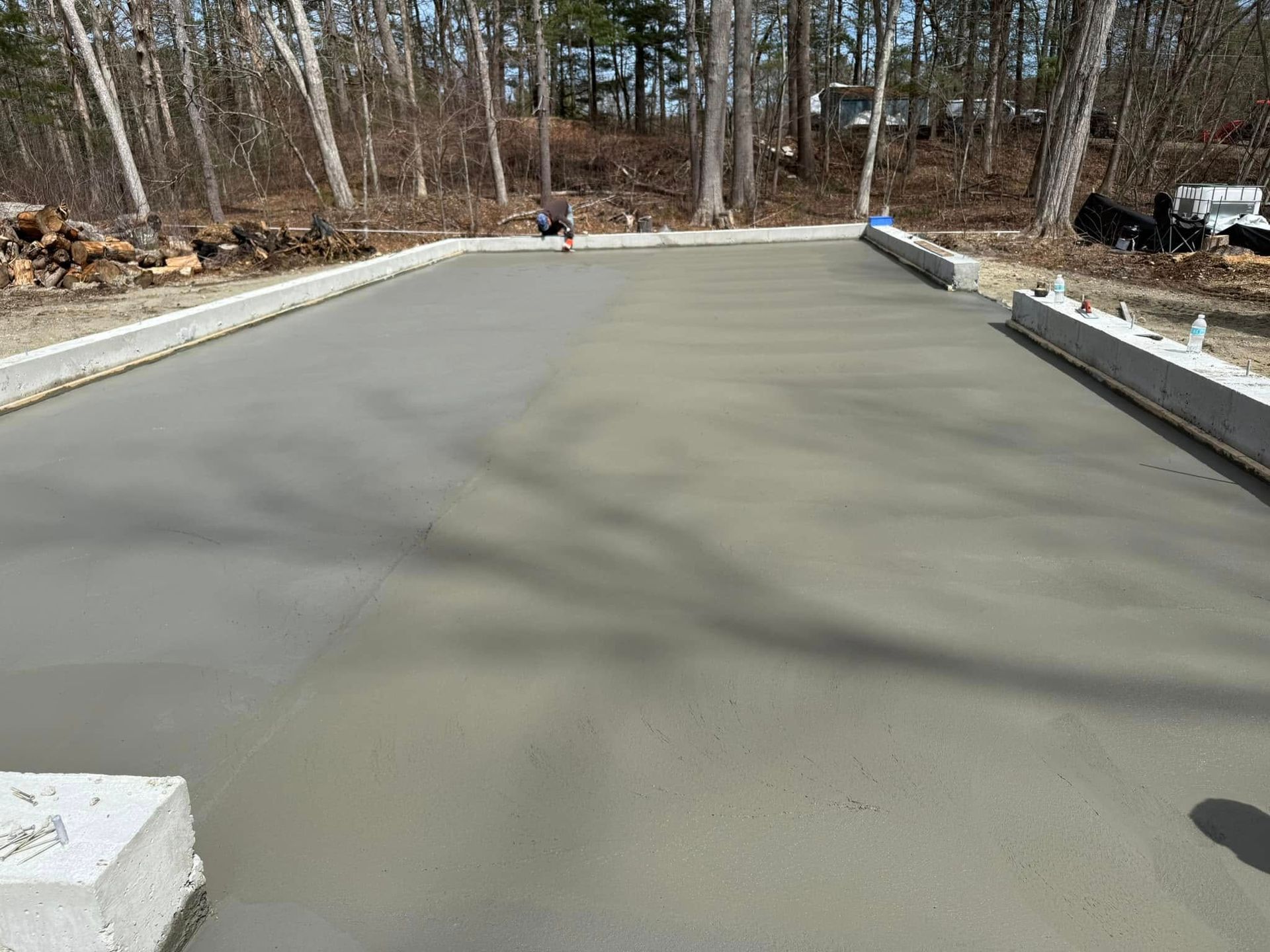 Freshly poured concrete surface with a person in the distance, bordered by concrete forms, in a wooded area.