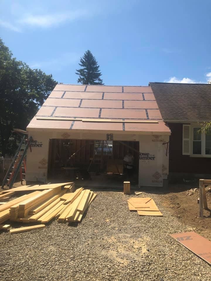 Garage addition under construction, exterior view; brown wood panels on roof, lumber on gravel driveway.