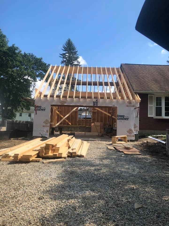 Garage under construction; wood frame, rafters visible, stacked lumber in foreground, house next to it.