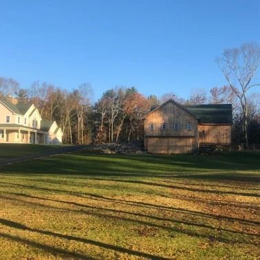 Farmhouse and barn on a sunny day with green lawn, trees, and blue sky.