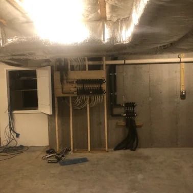 Electrical wiring installation in a basement; wooden frame, multiple wires, and conduit pipes against concrete wall.