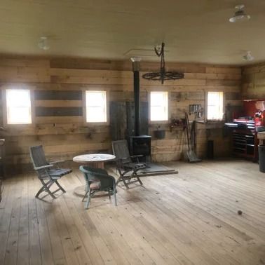 Wooden-walled interior with wood stove, three windows, two folding chairs, a small table, and a light-colored wooden floor.