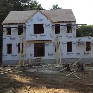 House under construction, covered in Tyvek wrap, with unfinished roof, windows, and wood.