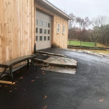 Exterior view of a wooden building with a garage door, asphalt driveway, and stone steps. A bench sits to the side.