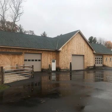 A light-colored wooden barn with several garage doors and a wet asphalt driveway under an overcast sky.